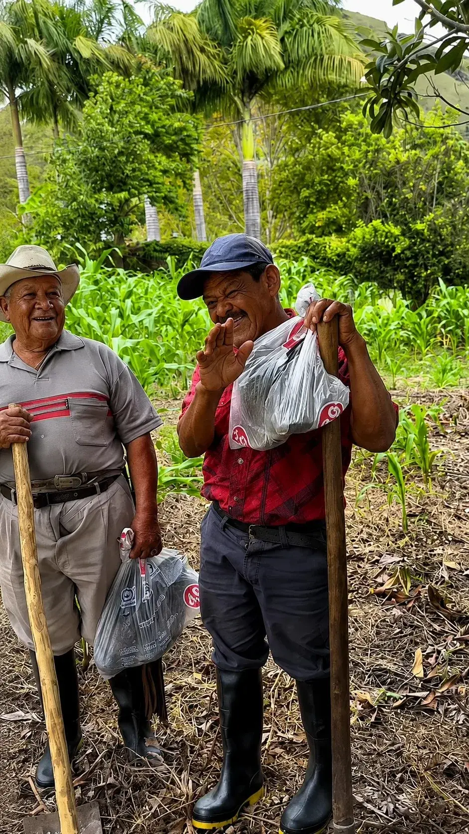 Two men standing in a field with farm tools, one holding bags of food