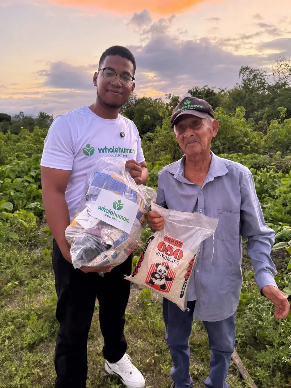 A Wholehuman team member handing a bag of food and a bag of rice to an older man in a field at sunset