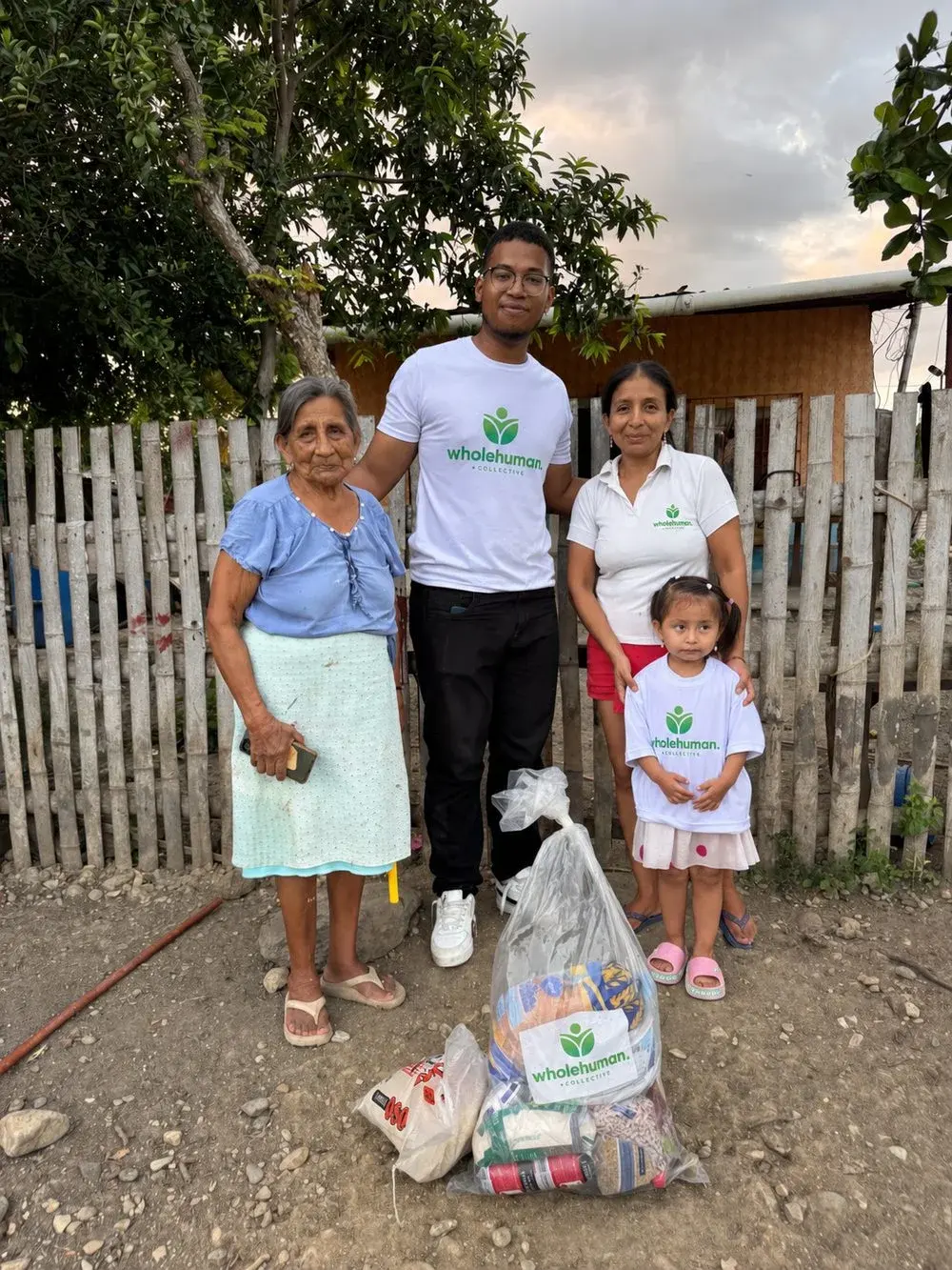 A Wholehuman team member with a grandmother, a mother and a little girl next to a bag of food outside their home